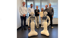 The image shows a group of six people in a bright office standing side by side behind two identical, white, humanoid robots. The two robots in the foreground feature a rounded base that widens at the bottom, slender bodies, articulated arms, and heads with large, pink-glowing eyes, along with a landscape-oriented tablet mounted on each of their chests. Behind them, standing from left to right, are four men and two women: a man in a light blue shirt and dark jeans, a man wearing glasses in a light blue shirt and dark pants, a woman with crossed arms in a black top and wide, light grey pants, a woman in a light beige jacket and white pants, a man in a dark shirt, and a man in a grey sweater. The group is standing in front of a large window with partially closed vertical blinds and a radiator underneath. The room has white walls and a dark, slightly speckled floor, with some cables visible on the ground in the right background.