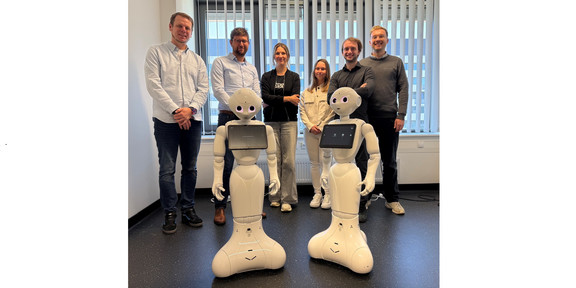 Das Bild zeigt eine Gruppe von sechs Personen in einem hellen Büro, die nebeneinander hinter zwei identischen, weißen, humanoiden Robotern stehen. Die beiden Roboter im Vordergrund haben eine abgerundete Basis, die nach unten hin breiter wird, schmale Körper, Gelenkarme und Köpfe mit großen, rosafarben leuchtenden Augen sowie jeweils ein querformatiges Tablet, das auf ihrer Brust montiert ist. Hinter ihnen reihen sich von links nach rechts vier Männer und zwei Frauen auf: ein Mann in hellblauem Hemd und dunkler Jeans, ein Brillenträger in hellblauem Hemd und dunkler Hose, eine Frau mit verschränkten Armen in schwarzem Oberteil und hellgrauer, weiter Hose, eine Frau in heller, beiger Jacke und weißer Hose, ein Mann in dunklem Hemd sowie ein Mann in grauem Pullover. Die Gruppe steht vor einer großen Fensterfront mit teilweise geschlossenen vertikalen Lamellenjalousien, unter der sich ein Heizkörper befindet. Der Raum hat weiße Wände und einen dunklen, leicht gesprenkelten Fußboden, auf dem auf der rechten Seite im Hintergrund einige Kabel liegen.