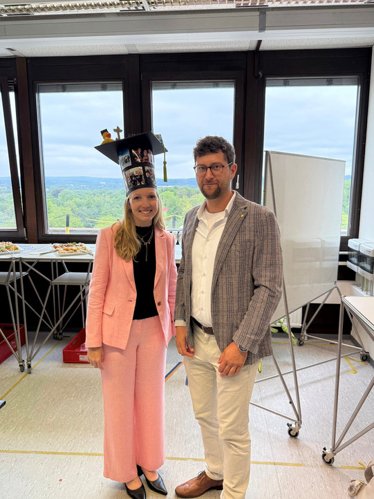 Das Bild zeigt eine Frau und einen Mann, die nebeneinander in einem hellen Raum stehen und in die Kamera lächeln. Die Frau auf der linken Seite hat langes blondes Haar und trägt einen hellrosa Hosenanzug mit einem schwarzen Oberteil darunter, eine goldene Halskette sowie schwarze, spitze Schuhe. Auf dem Kopf trägt sie einen auffälligen, individuell gestalteten Doktorhut. Der zylinderförmige Teil des Hutes ist mit mehreren Fotos beklebt, während auf der quadratischen Oberseite eine kleine Quietscheente mit eigenem Mini-Doktorhut und eine grüne Quaste befestigt sind. Rechts neben ihr steht ein Mann mit kurzen braunen Haaren, einem Bart und einer runden Brille. Er ist gekleidet in ein weißes Hemd, ein grau-schwarz gemustertes Sakko, eine helle beige Hose, einen dunklen Gürtel und braune Lederschuhe im Budapester-Muster; an seinem linken Handgelenk trägt er eine Armbanduhr. Im Hintergrund befinden sich große Fenster, die einen weiten Blick auf eine grüne, waldreiche Landschaft unter einem bewölkten Himmel bieten. Links im Raum steht ein hoher Tisch, auf dem Platten mit kleinen Snacks und Fingerfood angerichtet sind, auf dem Boden darunter befindet sich eine rote Plastikkiste. Auf der rechten Seite sind ein mobiles Whiteboard auf Rollen sowie Teile weiterer Büroeinrichtung zu erkennen. Der helle Fußboden ist an einigen Stellen mit gelben Klebebandstreifen markiert.