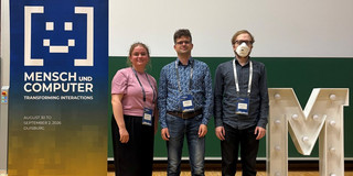 Drei Forschende stehen nebeneinander vor einer grünen Wand im Konferenzbereich der Mensch und Computer 2026. Links steht eine Frau in einem rosa T-Shirt, in der Mitte ein Mann mit Brille und gemustertem Hemd und rechts ein Mann mit Brille, FFP2-Maske und dunklem Hemd. Alle drei tragen Konferenz-Namensschilder. Links neben der Gruppe steht ein blaues Banner mit dem Konferenzmotto „Transforming Interactions“, rechts steht ein großer, weiß beleuchteter Buchstabe „M“.