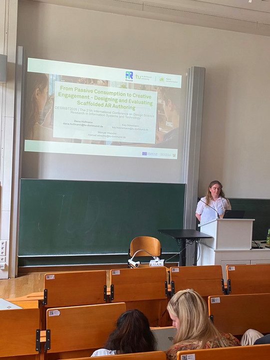 A shot from the perspective of an audience member in a lecture hall, showing a woman giving a presentation. She stands on the right side of the image at a white lectern, looking intently down at her laptop, wearing the same white eyelet top as seen in the group photo. Above her, a large presentation slide is projected onto a screen. The slide is titled "From Passive Consumption to Creative Engagement - Designing and Evaluating Scaffolded AR Authoring" and lists the names Dana Hofmann, Kay Hönemann, and Manuel Wiesche along with their email addresses below. Behind the speaker is a wide green chalkboard, and in front of it is a table holding a projector. In the foreground, the brown wooden seating rows of the lecture hall are visible, occupied by a few audience members whose backs and shoulders are out of focus.