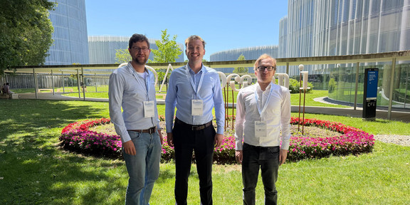The image shows three men standing side-by-side outdoors on a green lawn on a sunny day, looking directly at the camera. All three are wearing long-sleeved button-down shirts, long trousers, and lanyards with name badges clearly displaying the text "ECIS". The man on the left wears glasses, has a slight beard, and is dressed in a light blue shirt, blue jeans, and brown shoes. The man in the middle is smiling and wears a light blue shirt, dark trousers, a brown belt, and brown shoes. The man on the right wears glasses, a white shirt, dark trousers, and black shoes. Behind the men is a circular flower bed with an outer border densely planted with bright pinkish-red flowers. Directly behind the flower bed, large white sculptural letters stand on the grass, though they are partially obscured by the people. The background is dominated by modern buildings featuring striking, undulating white slatted facades that contrast against a clear, bright blue sky. A covered pedestrian walkway is visible in the background on the left, while a dark blue directional signpost stands on the right. In the bottom left corner of the foreground, a small sign is visible showing a red circle with a slash through a dog, indicating that dogs are not permitted.