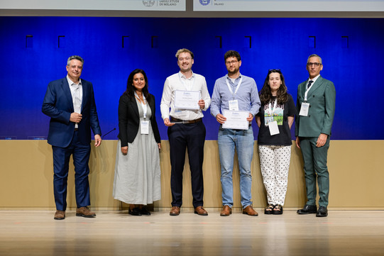 The image shows six people standing side-by-side on a light wooden stage in front of a long, beige-colored desk. In the background is a bright blue wall with vertical seams, above which is a white panel featuring partially visible logos, including the text "DEGLI STUDI DI MILANO". All six individuals are facing the camera and wearing white lanyards with name badges. From left to right: The first person wears a dark blue suit with a light-colored shirt open at the collar and brown shoes. The second person is dressed in a black blazer over a long, light grey dress and black flat shoes; a microphone is visible on the desk behind them. The third person wears a white button-up shirt, dark trousers, brown shoes, and glasses. This person is holding a framed certificate with the ECIS logo and a small glass trophy. The fourth person wears a blue and white striped shirt, blue jeans, brown shoes, and glasses, and is also holding a framed certificate. The fifth person is wearing a dark t-shirt with a colorful graphic print, wide-legged white trousers with black polka dots, and black platform sandals, with sunglasses perched on top of their head. The sixth person on the far right wears a sage green double-breasted suit, a white shirt, a dark tie, and black dress shoes.