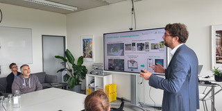 Das Foto im Querformat zeigt eine Präsentationssituation in einem hellen, modernen Seminar- oder Besprechungsraum. Auf der rechten Bildseite steht ein männlicher Referent im Profil, der den Blick nach links zu den Zuhörenden gerichtet hat. Er hat kurzes, lockiges braunes Haar, trägt eine Brille, ein blaues Sakko über einem weißen Hemd sowie eine blaue Jeans. Er gestikuliert mit beiden Händen, wobei er in der rechten Hand einen Presenter hält. Hinter ihm steht ein großer, digitaler Flachbildschirm auf einem mobilen Rollständer. Die darauf angezeigte Präsentationsfolie trägt die grüne Überschrift „Virtuelle Gärten – Einsatzmöglichkeiten“ und zeigt oben rechts das Logo der TU Dortmund. Der Inhalt der Folie ist in vier Bereiche unterteilt, die Bilder und Grafiken zu Augmented Reality (AR) und Virtual Reality (VR) Anwendungen auf Smartphones und Computerbildschirmen darstellen.  Auf der linken Bildhälfte sitzen die Zuhörenden an einer Anordnung aus weißen Tischen. Im direkten Vordergrund, mit dem Rücken zur Kamera, ist der Kopf einer Person mit rötlich-braunem, zum Dutt gebundenen Haar zu sehen, die einen schwarzen Pullover trägt. Dahinter sitzt ein Mann mit grau meliertem Haar und Bart in einem grauen Kapuzenpullover, der aufmerksam zum Referenten blickt. Vor ihm auf dem Tisch stehen eine gläserne Wasserkaraffe und mehrere Trinkgläser. Links im Hintergrund sitzt ein weiterer Mann mit Brille und blauem Hemd auf einem dunkelgrauen Sofa. Der Raum ist zweckmäßig eingerichtet: Im Hintergrund erkennt man ein weißes Regal, in dem silberne Koffer und gelbe Stapelboxen lagern, sowie eine große grüne Zimmerpflanze (Monstera) in der Ecke. An den weißen Wänden hängen gerahmte Bilder, und an der Decke sind längliche Leuchtstoffröhren angebracht.  English The landscape-format photo captures a presentation taking place in a bright, modern seminar or meeting room. On the right side of the image, a male presenter stands in profile, facing left towards the audience. He has short, curly brown hair, wears glasses, a blue blazer over a white shirt, and blue jeans. He is gesturing with both hands whilst holding a presentation clicker in his right hand. Behind him is a large digital flat-screen display mounted on a rolling stand. The slide being presented features the green headline "Virtuelle Gärten – Einsatzmöglichkeiten" (Virtual Gardens – Application Possibilities) and shows the logo of TU Dortmund in the top right corner. The slide content is divided into four quadrants displaying images and graphics related to Augmented Reality (AR) and Virtual Reality (VR) applications on smartphones and computer monitors.  On the left side of the image, the audience is seated at a configuration of white tables. In the immediate foreground, with their back to the camera, is the head of a person with reddish-brown hair tied in a bun, wearing a black sweater. Further back sits a man with grey-flecked hair and a beard, wearing a grey hoodie, who is looking attentively at the speaker. A glass water carafe and several glasses are placed on the table in front of him. In the background on the far left, another man wearing glasses and a blue shirt is seated on a dark grey sofa. The room is functionally furnished: in the background, a white shelving unit holding silver cases and yellow stacking bins is visible, alongside a large green houseplant (Monstera) in the corner. Framed pictures hang on the white walls, and long fluorescent light fixtures are mounted on the ceiling.