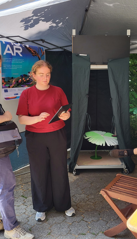 A young woman in a red t-shirt stands under a canopy on a sunny day, looking at a tablet in her hands. Behind her hangs a poster with the text "flow AR". Right next to it is a dark, open booth. Inside the booth, an illuminated, flat flower-shaped object lies on the floor.