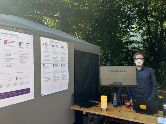A young man wearing glasses and an FFP2 mask stands outdoors behind a wooden table. On the table are a monitor displaying the website "DEIN DATENSPIEGEL", two open laptops, drinks, and a glowing orange cylindrical lamp. To the left of the stand is a grey partition board with two large posters about smart home privacy.