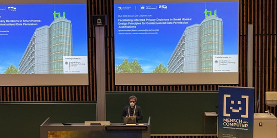 Ein Vortragender mit Brille, FFP2-Maske und dunkler Kleidung spricht an einem Rednerpult auf der Konferenz Mensch und Computer 2026. Auf den Projektionsleinwänden ist die Titelfolie seines Vortrags über Gestaltungsprinzipien für Privatsphäre-Entscheidungen im Smart Home eingeblendet. Die Folie zeigt das Logo der TU Dortmund, die Autorennamen Björn Konopka und Manuel Wiesche sowie eine Fotografie des TU-Dortmund-Matheturms vor blauem Himmel.
