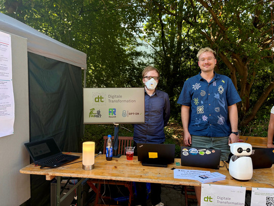 Two men stand outdoors behind a wooden information booth. The man on the left wears an FFP2 mask; the man on the right wears a dark blue shirt with light patterns. A monitor in the center of the table displays the "dt Digitale Transformation" logo and three smaller project logos. The table also holds two laptops, an orange lamp, informational flyers, and a small white humanoid robot.