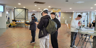 Das Foto im Querformat zeigt eine belebte Szene in einem geräumigen, gut beleuchteten Foyer einer öffentlichen Einrichtung, vermutlich einer Universität oder Schule. Der Boden ist auffällig mit rotbraunen, sechseckigen Fliesen gemustert. Im Zentrum der Handlung stehen zwei Personen, die weiße Virtual-Reality-Headsets tragen und Handcontroller bedienen. Sie befinden sich innerhalb eines auf dem Boden mit lila Klebeband markierten quadratischen Bereichs. Innerhalb dieses Quadrats bilden Streifen aus grünem Klebeband die großen Buchstaben „VR“. Die Person im direkten Vordergrund trägt eine dunkle Jacke, hellgraue Jeans und einen olivgrünen Rucksack auf dem Rücken. Links daneben steht die zweite Person mit VR-Brille, ganz in Dunkelblau und Schwarz gekleidet. Ein dritter Mann in schwarzer Kleidung steht rechts neben der VR-Zone und beobachtet die Nutzer. Weiter rechts im Bild steht ein junger Mann mit hellem Haar, bekleidet mit einem weißen Zopfmuster-Pullover und grauen Jeans, an einem hohen runden Stehtisch. Er blickt auf Unterlagen oder ein Gerät hinab; vor ihm steht ein Pappbecher. Im Hintergrund links ist eine Glasstruktur mit Metallrahmen zu sehen, sowie Tische mit Laptops und gelben Stühlen. Eine runde Säule in der Mitte ist mit diversen Zetteln und Flyern plakatiert. Im weiteren Hintergrund erkennt man unverputzte Ziegelwände, große Fensterfronten und sitzende Personen. Die Decke ist weiß und mit eingelassenen runden Leuchten versehen.