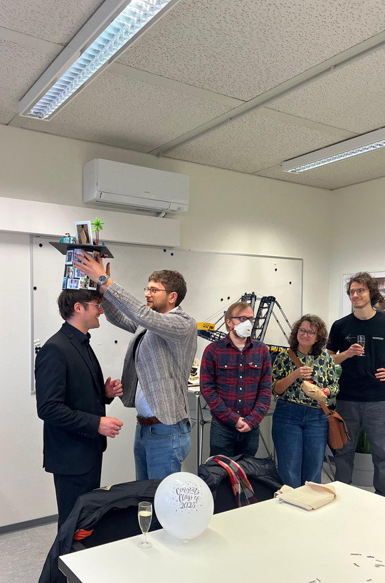 The photo captures a celebration in a brightly lit office or seminar room, featuring a group of five people, likely marking an academic milestone like a PhD defense. On the left side, the main focus is on a smiling man dressed in a black suit and black shirt. He is standing still while another man places a custom-made graduation cap (mortarboard) on his head. The man placing the hat wears a grey plaid blazer, blue jeans, and glasses. The hat itself is heavily decorated; the cylindrical part is covered in small printed photos, and the top features miniature objects, including a small palm tree and a frame.  To the right of this pair, three onlookers observe the scene. Closest to the center is a man wearing a red plaid shirt, jeans, and glasses, who is also wearing a white face mask and has his hands clasped. Next to him stands a woman with curly hair and glasses, wearing a yellow and green patterned sweater; she holds a glass of sparkling wine and a brown bag. On the far right stands a tall man in a black t-shirt and grey trousers, also holding a glass of sparkling wine and smiling.  In the immediate foreground, resting on a white table, is a white balloon with the text "Congrats Class of 2025" printed in a festive font. The background features a whiteboard, a wall-mounted air conditioning unit, and a display area featuring a large model of a construction crane or excavator (likely a Lego Technic model). The room has a standard drop ceiling with fluorescent lighting.
