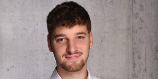 The image displays a professional portrait of a young man positioned in front of a light grey, textured concrete wall. He has a fair complexion, dark brown eyes, and dark brown hair that is faded on the sides and styled with a slight wave on top. He sports a neatly trimmed beard concentrating around the mouth and chin, with light stubble on his cheeks. The man is wearing a crisp white button-down shirt with the collar open at the top, creating a smart-casual look. His torso is angled slightly to his right, while his face is turned directly toward the camera. He maintains direct eye contact with the viewer, offering a subtle, friendly, and relaxed smile with closed lips.
