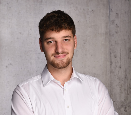 Das Bild ist ein professionelles Porträt eines jungen Mannes vor einer hellgrauen, strukturierten Betonwand. Er hat einen hellen Hautton, dunkelbraune Augen und dunkelbraunes Haar, das an den Seiten kurz geschnitten ist und am Oberkopf länger sowie leicht gewellt fällt. Er trägt einen gepflegten, kurz getrimmten Bart, der sich um den Mund und das Kinn zieht, sowie leichte Stoppeln an den Wangen. Der Mann ist mit einem weißen Langarmhemd bekleidet, dessen oberster Knopf geöffnet ist, was einen legeren, aber ordentlichen Eindruck vermittelt. Sein Oberkörper ist leicht nach rechts gedreht, während sein Kopf frontal zur Kamera ausgerichtet ist. Er blickt den Betrachter direkt an und zeigt ein geschlossenes, freundliches und entspanntes Lächeln.