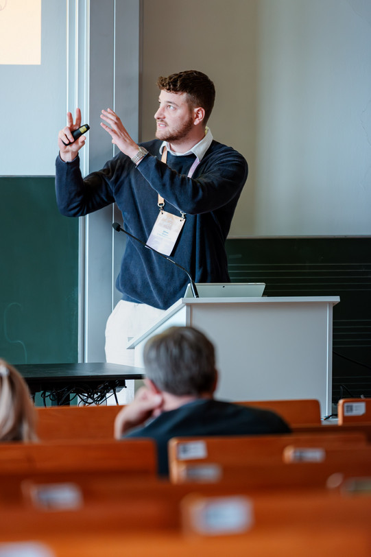 A photograph taken from the audience's perspective in a brightly lit lecture hall. In the center, a man with dark curly hair and a beard stands at a white lectern. He is wearing a dark blue ribbed sweater over a white shirt, light trousers, and a lanyard with a conference name badge. He is looking to the left, over the audience, and gesturing animatedly with both hands raised, holding a black presentation clicker in his right hand. A laptop and a gooseneck microphone are on the simple lectern. In the blurred foreground, the backs of two audience members and rows of brown wooden benches are visible. In the background, there is a green chalkboard and a brightly lit white projection screen.