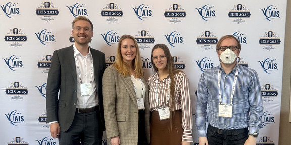 The image is a full-body group photo showing four people standing side-by-side facing the camera in front of a conference backdrop. The backdrop is white and features a banner at the top reading "ICIS 2025" in large dark blue letters, followed by "Nashville Tennessee December 14 – 17". The banner includes graphics of a guitar headstock on the left and an orange guitar neck on the right containing the hashtag #ICIS2025. Below the banner, the wall features a repeating step-and-repeat pattern of logos for "AIS" (Association for Information Systems) and "ICIS 2025".  From left to right: The first person is a man with short blond hair and glasses, wearing a dark grey suit jacket, a white button-down shirt, dark trousers, and brown leather dress shoes. Next to him is a woman with long blond hair wearing a beige checked blazer, a white top, black trousers, and light-colored sneakers. Beside her stands a woman with long brown hair and glasses, wearing a vertically striped shirt in white and reddish-brown, a brown midi skirt, dark tights, and tan boots with fur trim. On the far right is a man with short hair and glasses wearing a white face mask. He is dressed in a blue checkered button-down shirt, dark jeans, and brown boots. All four individuals are wearing conference name badges on lanyards around their necks. They are standing on a carpet patterned with large, abstract leaf shapes in shades of blue and green.